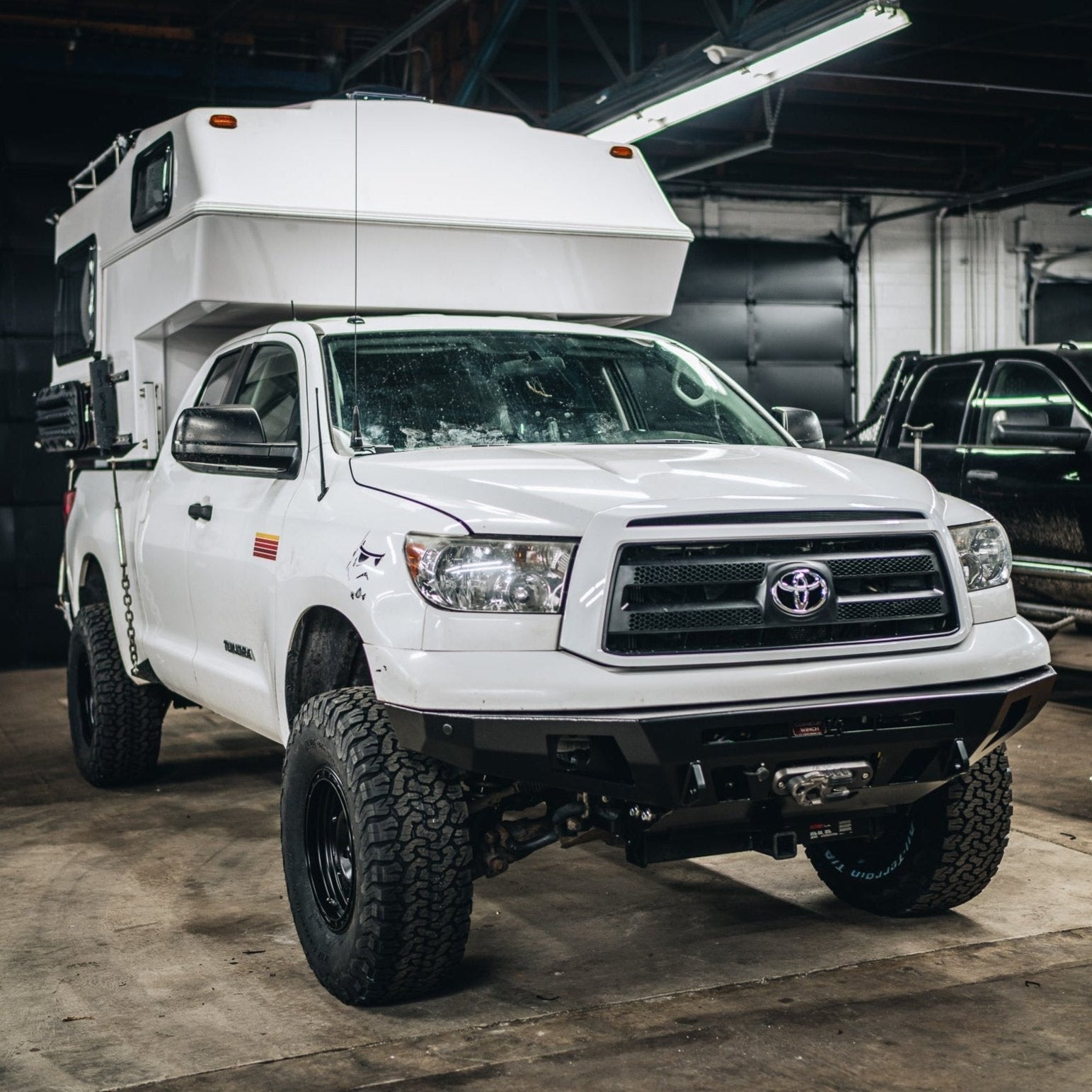 C4 Overland plate bumper on a lifted and modified white Toyota Tundra with a fiberglass slide-in camper installed.